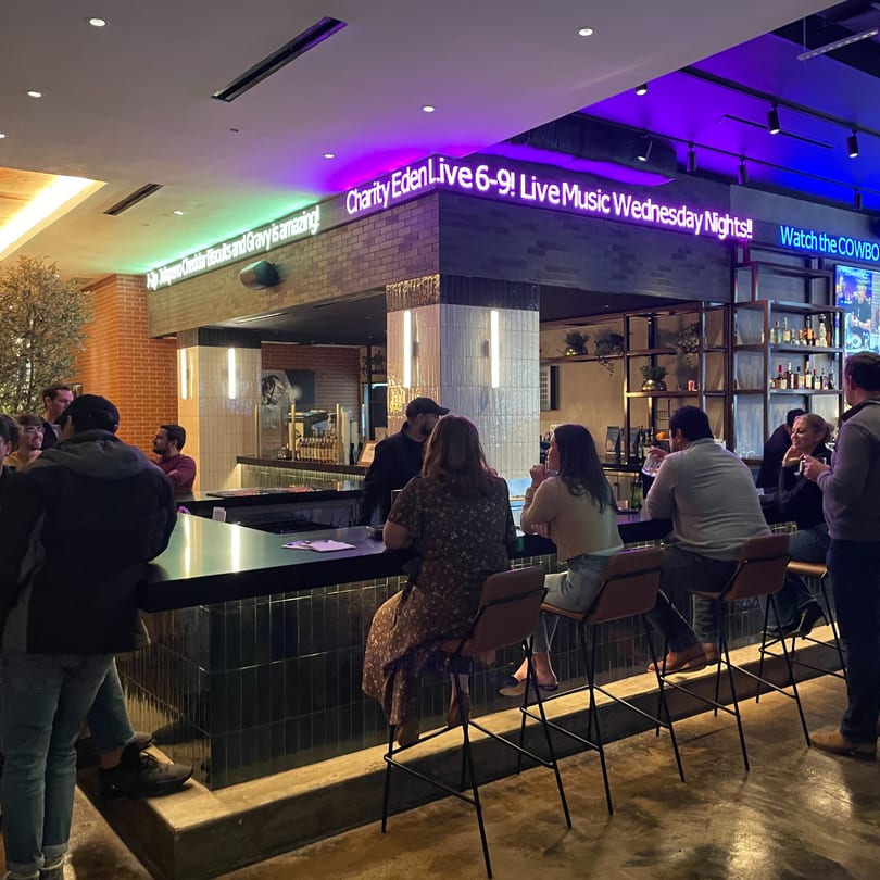 Patrons enjoying drinks at The Operators Club bar with neon signs advertising live music and food.