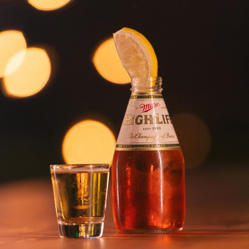 Miller High Life bottle with a lemon wedge and a shot glass on a table with bokeh lights.