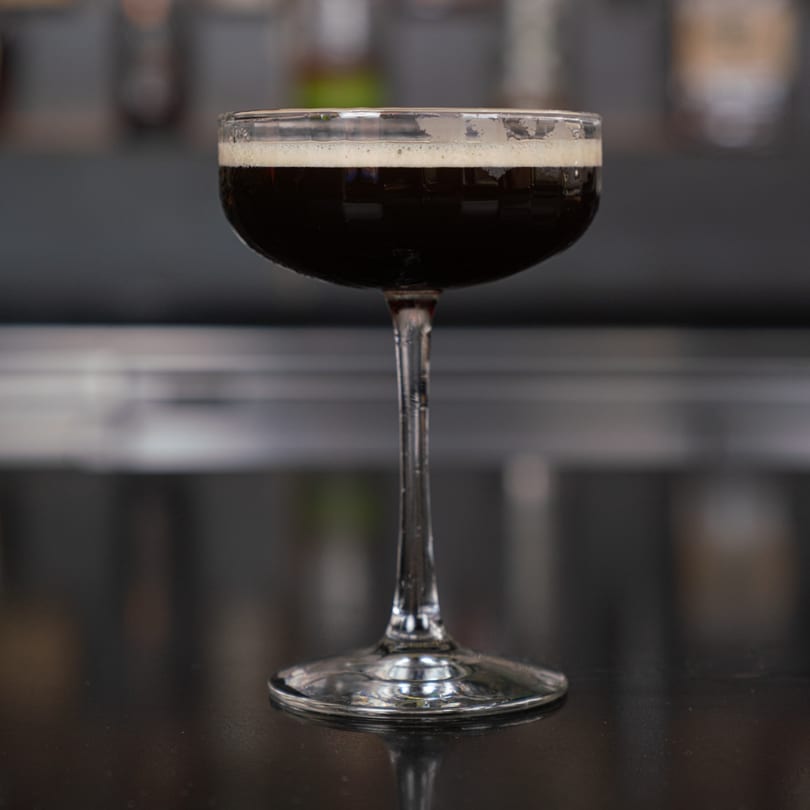 A dark cocktail with a foamy top in a coupe glass on a dark, reflective surface.