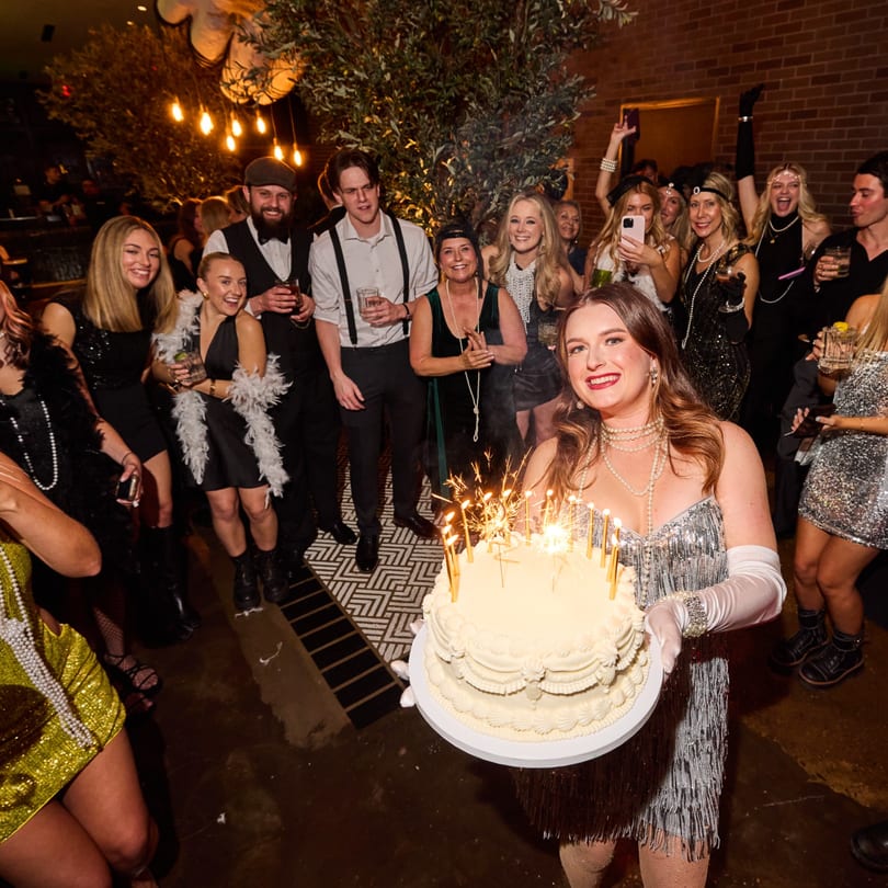 A woman in a flapper dress holds a birthday cake with sparklers, surrounded by smiling guests.