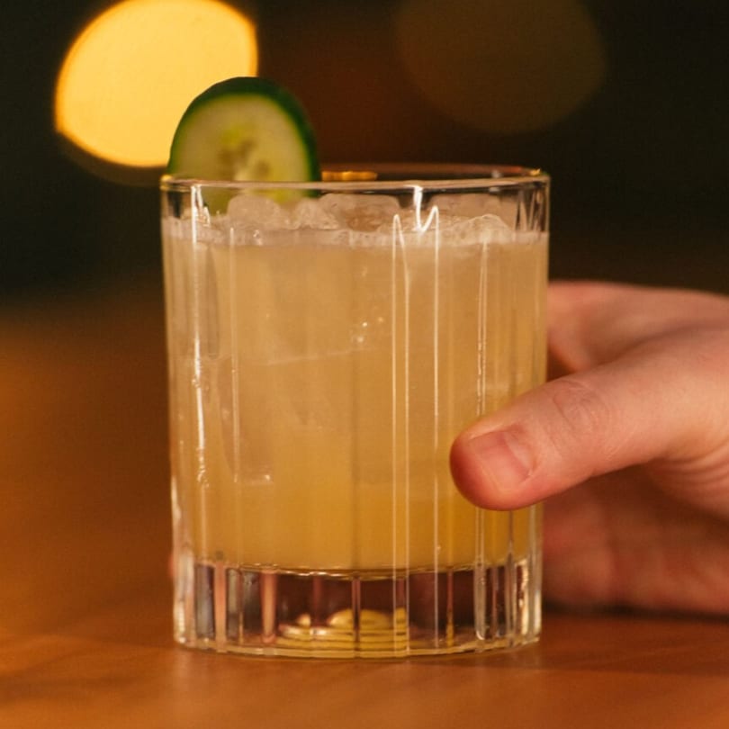 Hand holding a cocktail with a cucumber slice garnish, against a blurred, warm-lit background.