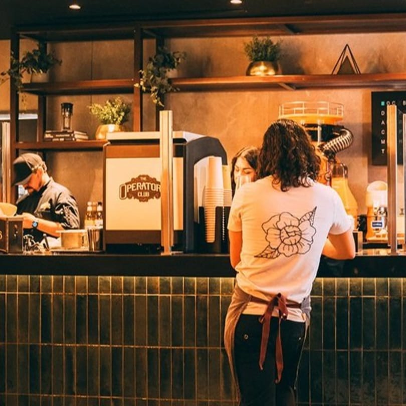 Baristas at The Operators Club preparing drinks behind a dark tiled counter with shelves.