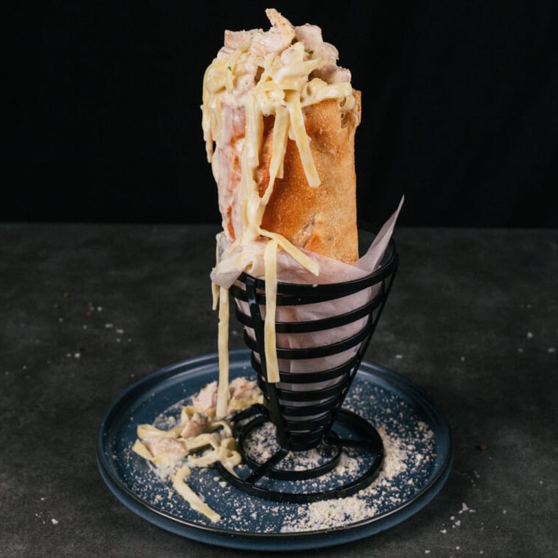 A baguette filled with creamy chicken and cheese, served upright in a black metal holder on a blue plate.