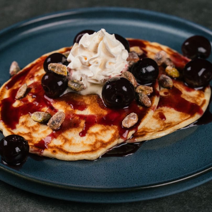 Two pancakes topped with whipped cream, cherries, candied nuts, and berry syrup on a blue plate.