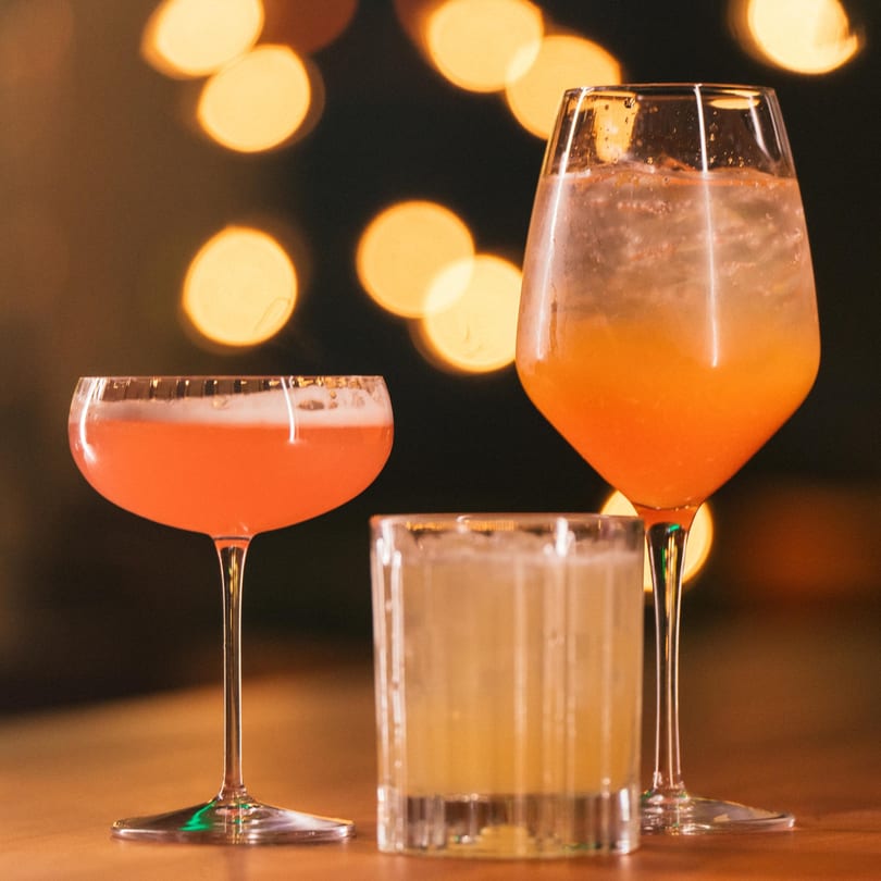 Three cocktails, one pink, one orange, and one yellow, on a bar with bokeh lights.