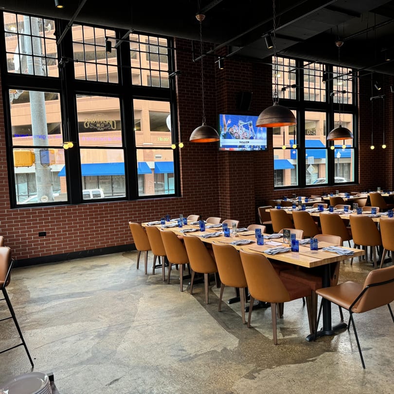 The Operators Club dining room with long wooden tables, brown chairs, brick walls, and large windows.