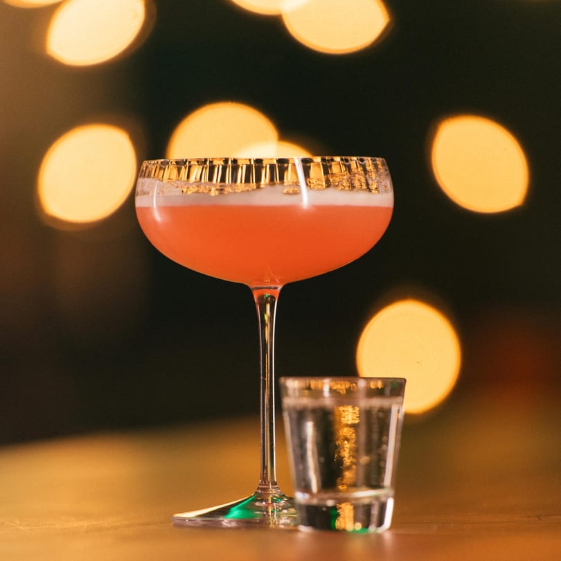A vibrant pink cocktail with a golden rim and a shot glass on a warm, blurry background.