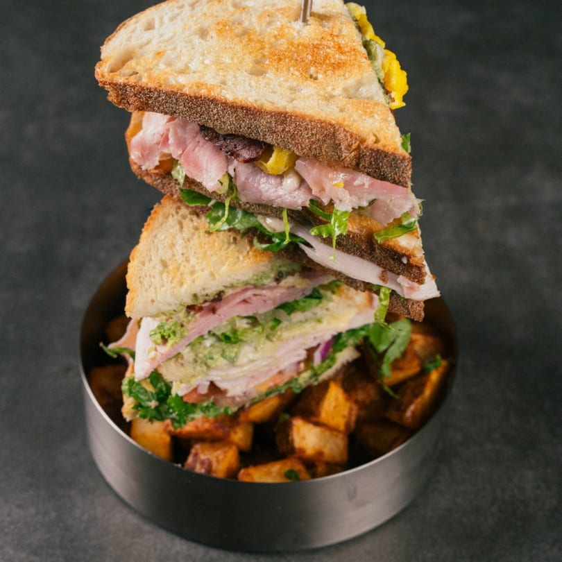 A stacked sandwich with ham, lettuce, and cheese, served with roasted potatoes in a metal bowl.