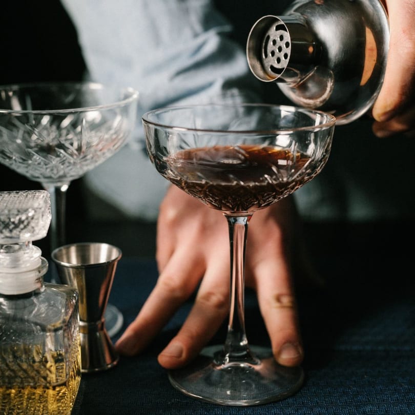 A bartender pours a dark cocktail from a shaker into a crystal coupe glass at The Operators Club.