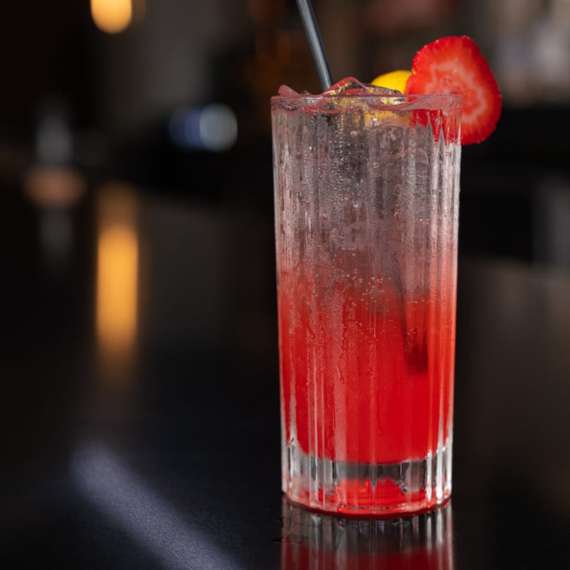 A vibrant red cocktail in a tall glass, garnished with a strawberry slice and lemon, on a dark bar.