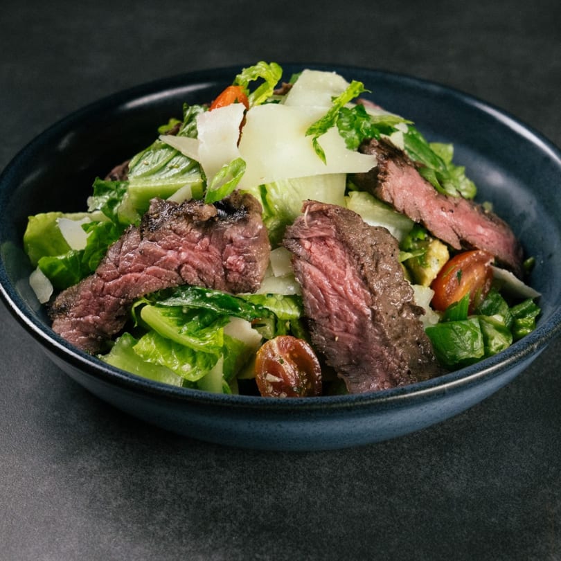 The Operators Club's steak salad with romaine, cherry tomatoes, avocado, and shaved parmesan in a dark bowl.