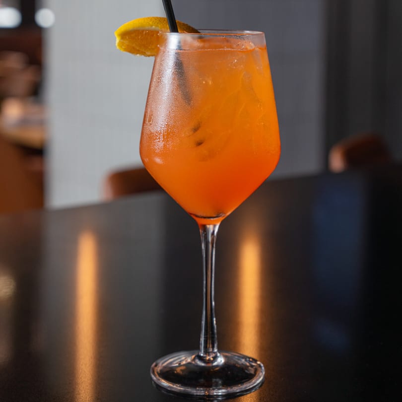 A vibrant orange cocktail in a wine glass, garnished with an orange slice and a black straw.