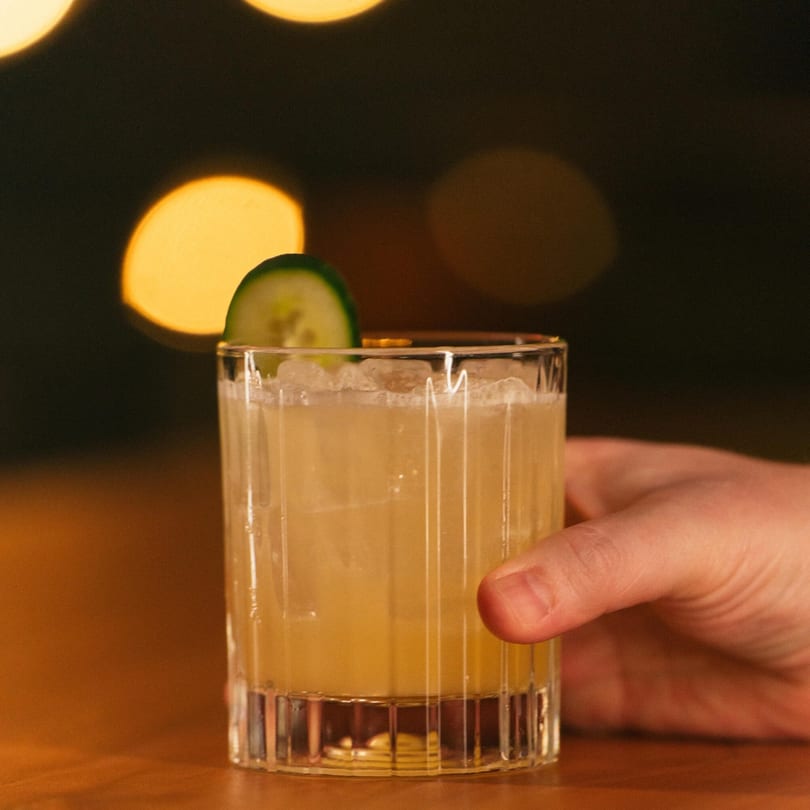 A hand holding a cocktail garnished with a cucumber slice, against a dark, bokeh background.