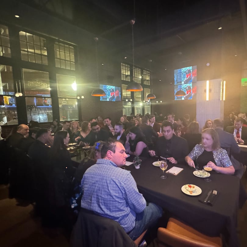 Diners seated at long tables in a dimly lit restaurant with large windows and hanging lights.