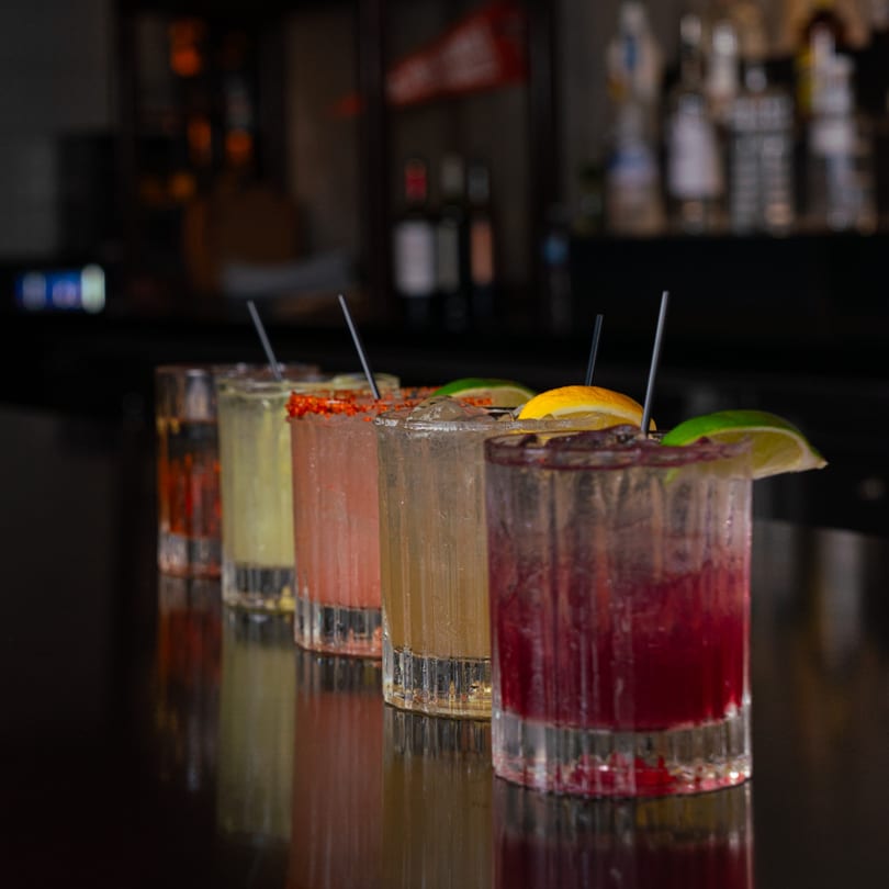 Five colorful cocktails with garnishes and straws lined up on a dark bar at The Operators Club.