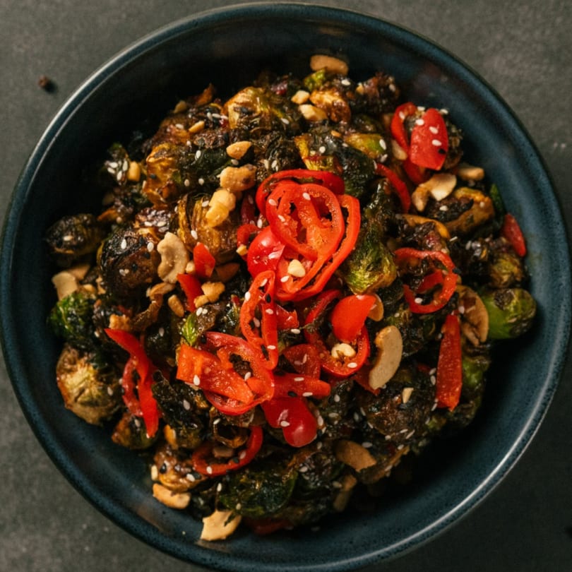 Roasted Brussels sprouts with red peppers, cashews, and sesame seeds in a dark blue bowl.