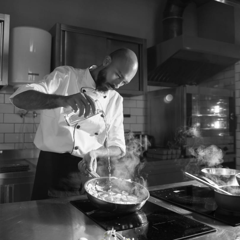 A chef in a white coat pours liquid from a glass into a steaming pan of food.