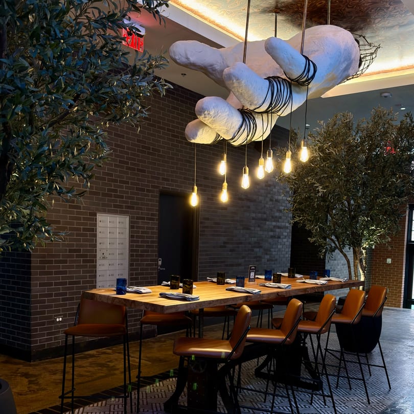 A long wooden communal table with brown bar stools under a large hand sculpture and Edison bulb lights.